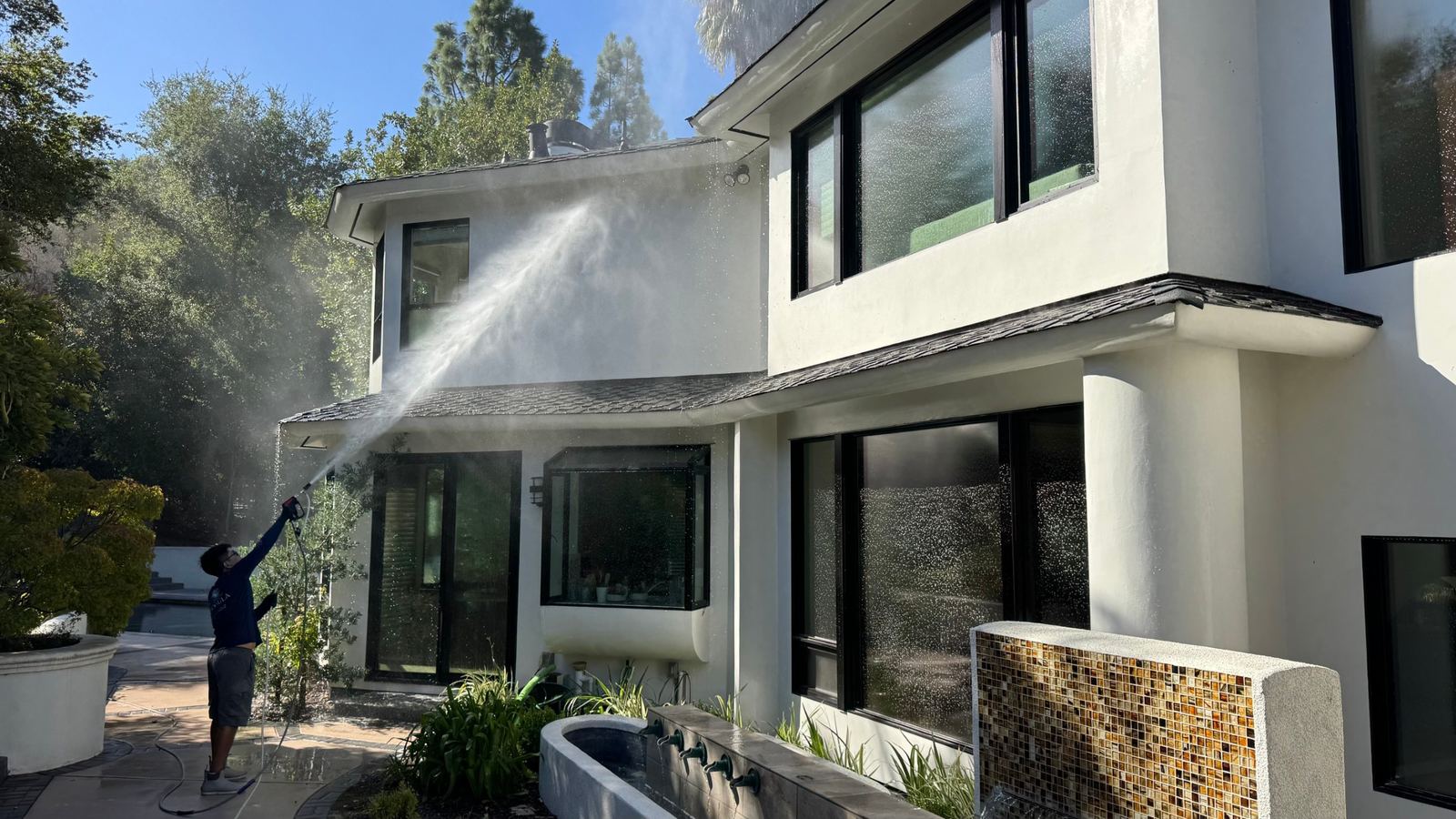 Technician soft washing the upper walls and eaves of a white two-story stucco home with a low-pressure spray